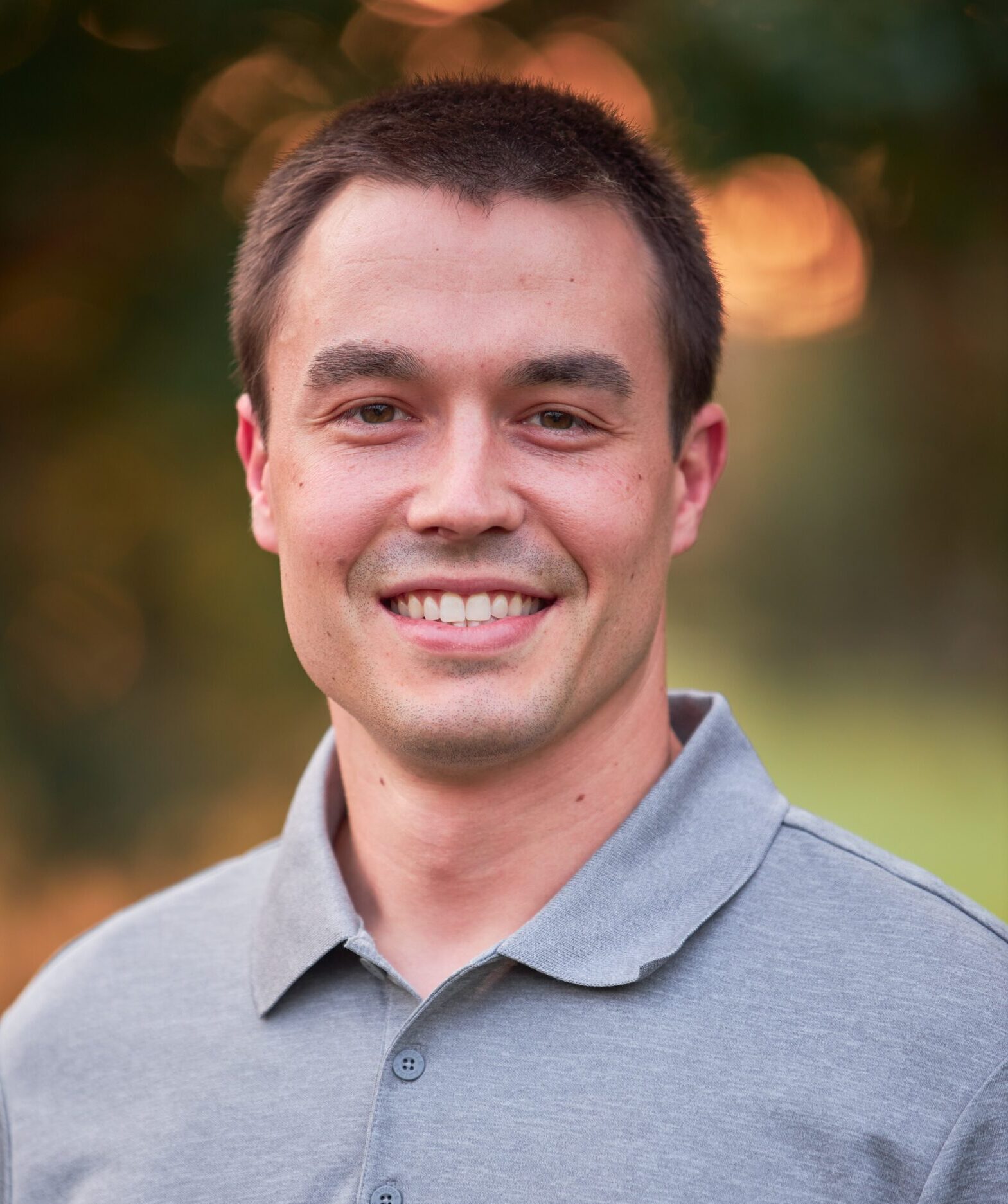 Keaton Dugan, with short dark hair and a gray polo shirt, smiles warmly at the camera against a blurred natural background.
