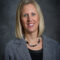 A woman with shoulder-length blonde hair is wearing a gray blazer and black top, smiling in front of a dark gray background. Her confident demeanor suggests she might be Kerry Hatzenbuehler.