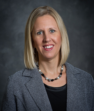 A woman with shoulder-length blonde hair is wearing a gray blazer and black top, smiling in front of a dark gray background. Her confident demeanor suggests she might be Kerry Hatzenbuehler.