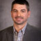 Kody Winterscheidt, in a gray suit and checkered shirt, smiles warmly against the muted gray background.