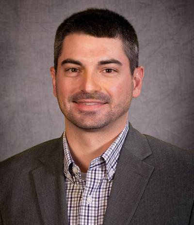 Kody Winterscheidt, in a gray suit and checkered shirt, smiles warmly against the muted gray background.