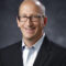 Brian Kuehl, a bald man with glasses, smiles warmly while donning a dark suit jacket and light shirt against a dark, blurred background.