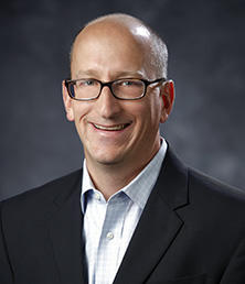 Brian Kuehl, a bald man with glasses, smiles warmly while donning a dark suit jacket and light shirt against a dark, blurred background.