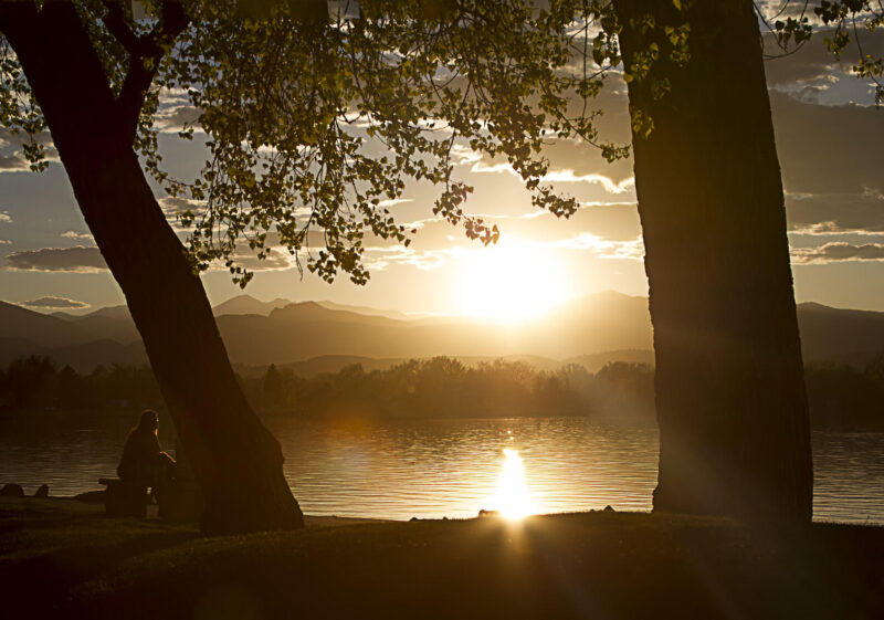 Sunset over a lake, framed by two large trees. Silhouette of a person sitting on a bench in the foreground. Mountains and scattered clouds visible in the background.