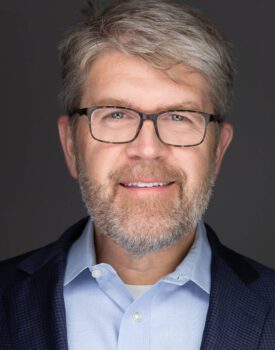 Lance Woodbury, a bearded man with glasses, smiles in a professional headshot, donning a blue shirt and dark jacket against a plain gray background.