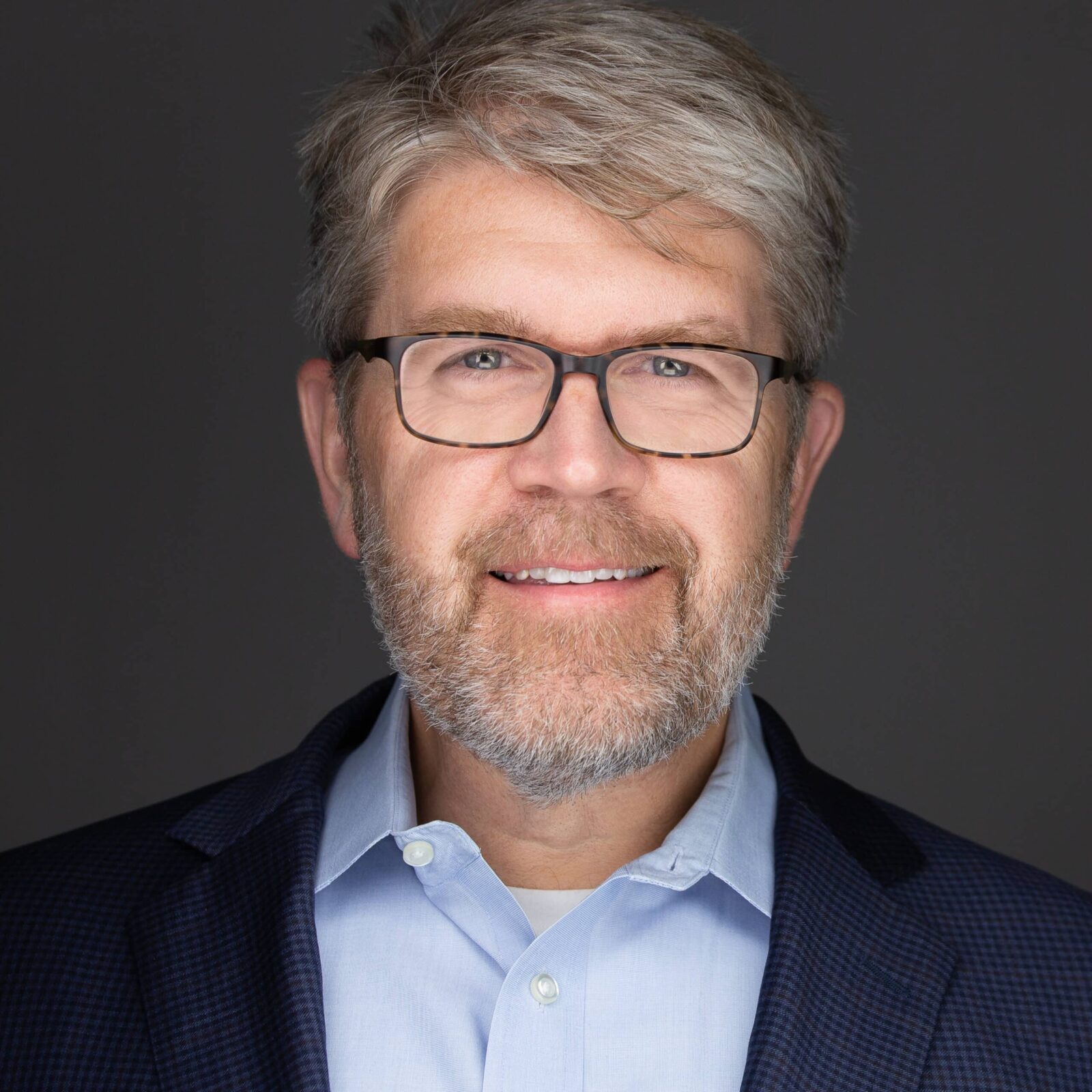 Lance Woodbury, a bearded man with glasses, smiles in a professional headshot, donning a blue shirt and dark jacket against a plain gray background.