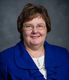 A person with short brown hair and glasses, resembling Corlene Lange, is wearing a blue blazer and white top, smiling warmly against a gray background.