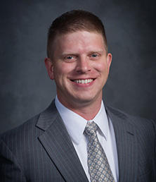Travis Lank, dressed in a sharp suit and tie, radiates confidence with a warm smile against the dark background.