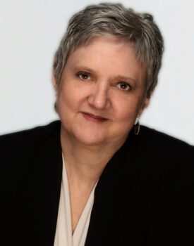 Laura Sands, with her short grey hair, dons a black blazer over a light blouse, her smile gentle against the plain backdrop.