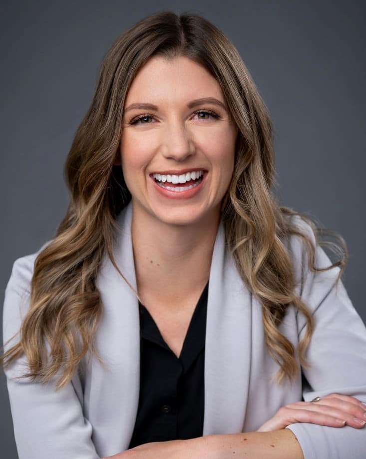 Lisa Becker, a smiling person with long wavy hair, dons a light gray blazer over a black top, set against a dark gray background.