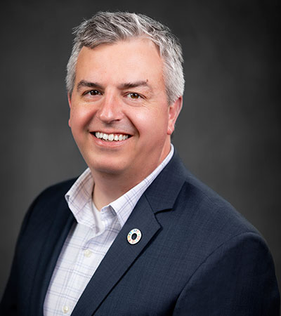 Smiling man with gray hair in a dark suit and white shirt, wearing a circular pin, against a dark background, exuding an air of confidence reminiscent of Matthew Armstrong.