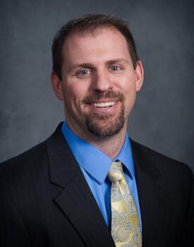 Maxson Irsik, a man with short hair and a beard, wears a black suit, blue shirt, and patterned yellow tie, smiling confidently against a gray background.