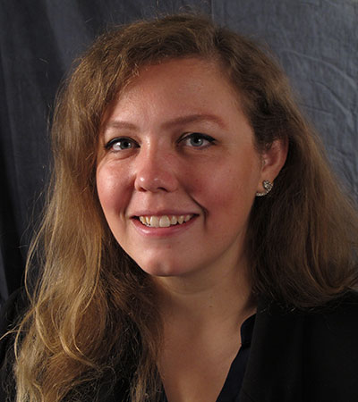 A person with long, wavy hair and a black top smiles at the camera against a dark background.