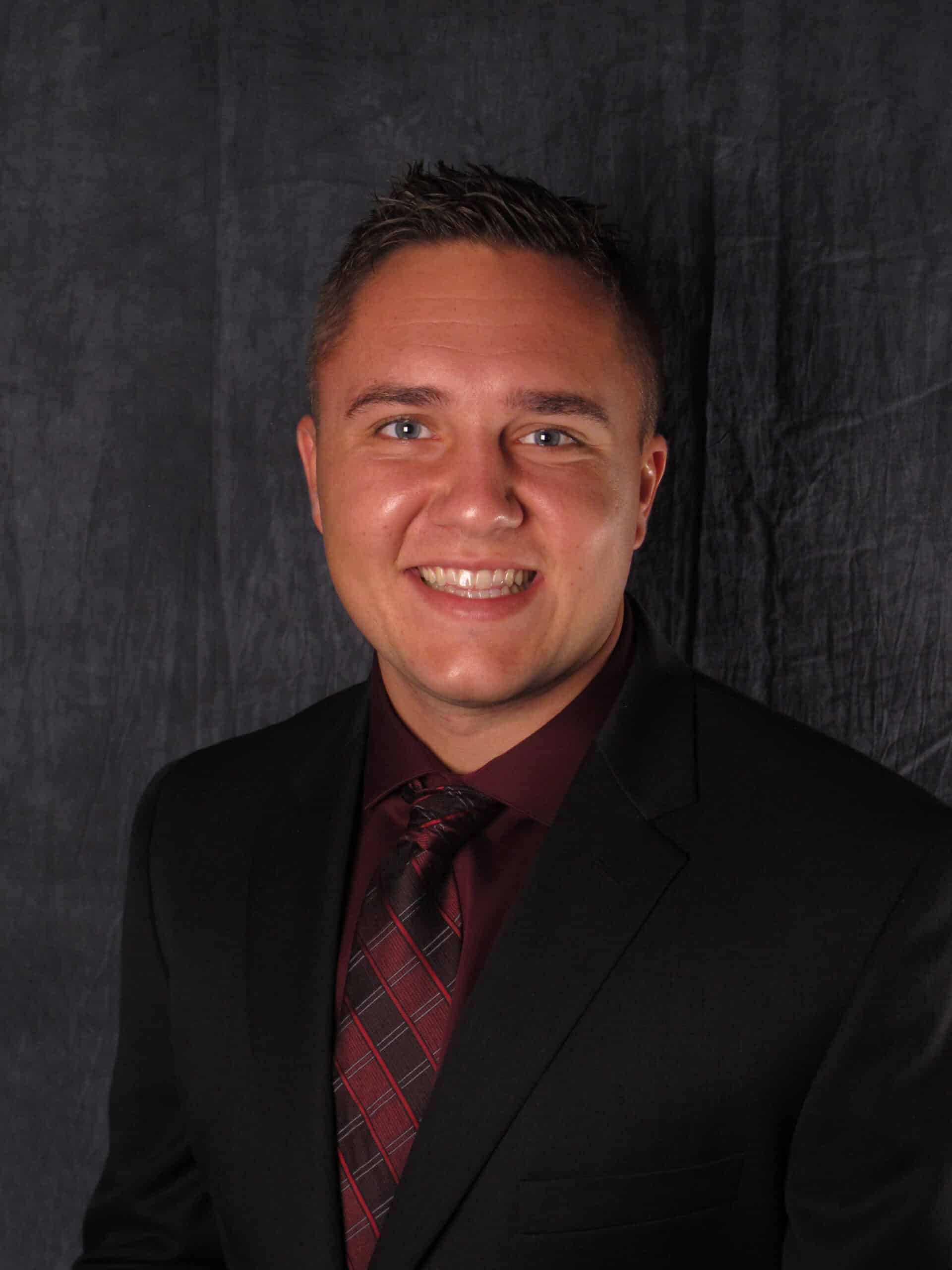 Michael Swift, in a suit and tie, smiles confidently in a professional studio portrait against a dark background.