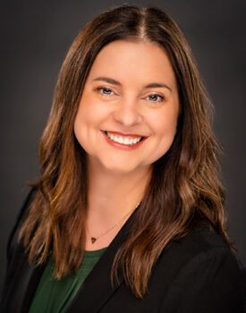 Michelle Ellis, with her long brown hair and a warm smile, stands against a dark background, wearing a dark blazer over a green top.