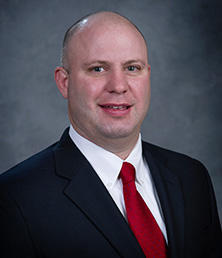 Kevin Mills, a bald man in a suit and red tie, poses with a neutral expression against a gray background.