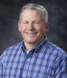 Doug Mitchell beams in a blue plaid shirt against a dark, textured background.
