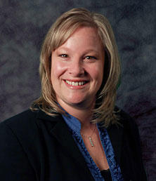 Christy Norton, with shoulder-length blonde hair, beams a bright smile while wearing a dark blazer and blue shirt against a textured gray background.