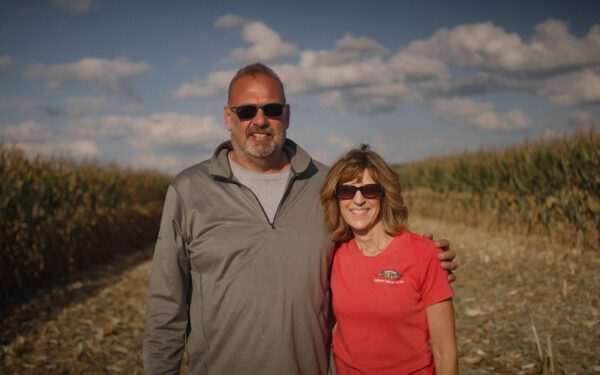 Two people wearing sunglasses stand together in a cornfield under a partly cloudy sky, smiling at the camera—celebrating moments in Food and Ag Production.