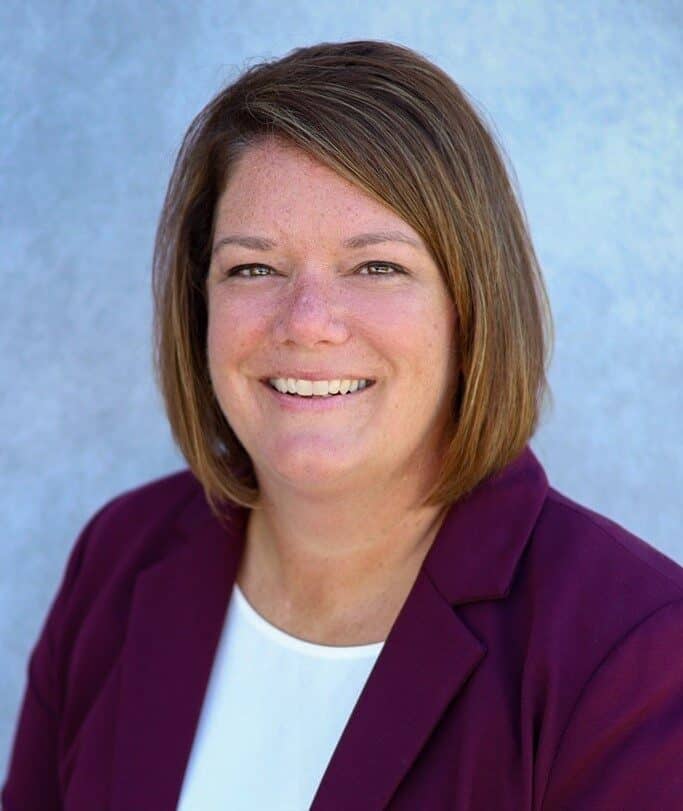 Jena Petersen, with short brown hair, is smiling in a maroon blazer and white shirt against a light blue background.
