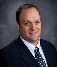 Chris Pfannenstiel, dressed in a sharp suit and tie, smiles confidently against a gray background.