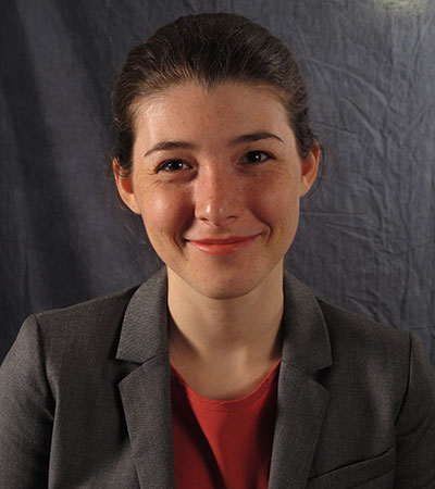 Person smiling in a professional portrait, wearing a gray blazer over a red top, against a plain dark backdrop.