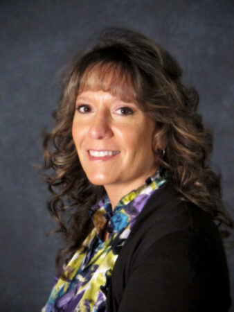 A woman with wavy hair smiles while wearing a floral blouse and dark sweater, set against a dark background.
