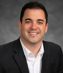Austin Sterling, in a dark suit and white shirt, smiles at the camera against a gray background.