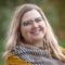 Elizabeth Swanson, sporting glasses and a warm smile, enjoys the outdoors. Her shoulder-length hair flows gently as she wears a multicolored scarf. The background is beautifully blurred with green and brown tones.