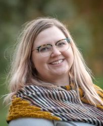 Elizabeth Swanson, sporting glasses and a warm smile, enjoys the outdoors. Her shoulder-length hair flows gently as she wears a multicolored scarf. The background is beautifully blurred with green and brown tones.