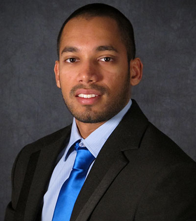 Tharin Wanigasekara, elegantly attired in a suit and blue tie, poses confidently against a dark background, his short hair neatly styled and beard precisely trimmed.