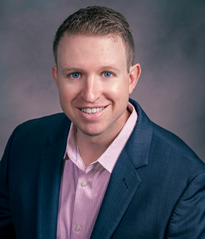 Travis Free, wearing a blue suit and pink shirt, smiles warmly at the camera against a gray background.