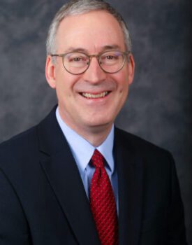 Loren Young, a smiling man with glasses, is elegantly dressed in a dark suit, light blue shirt, and red tie as he poses confidently against a dark gray backdrop.