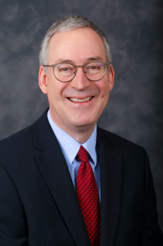 Loren Young, a smiling man with glasses, is elegantly dressed in a dark suit, light blue shirt, and red tie as he poses confidently against a dark gray backdrop.