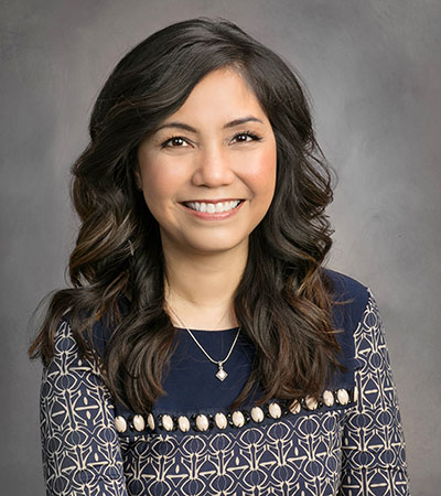 Amanda Huffman, with her long dark hair and a bright smile, wears a patterned navy dress against a plain gray background.