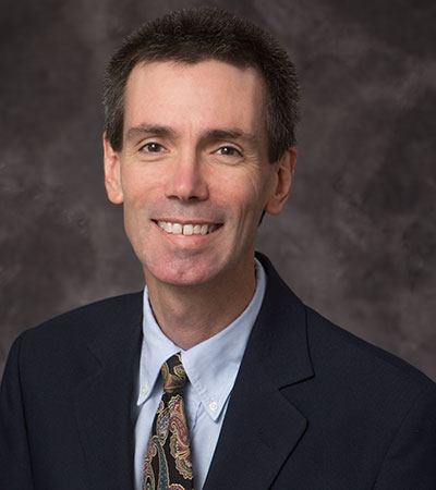 A smiling man in a suit with a patterned tie stands confidently against a dark background, capturing the charisma reminiscent of Bill Woolsey.