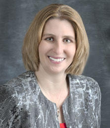 Becky Sweet, with her shoulder-length hair and patterned jacket, flashes a warm smile against a neutral background.