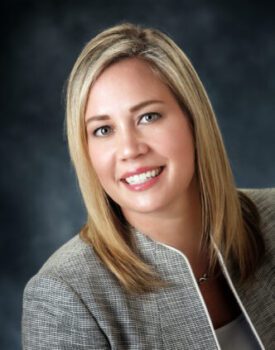 Jen Hertzig, with shoulder-length blonde hair, wearing a gray blazer and white top, smiles at the camera against a dark background.