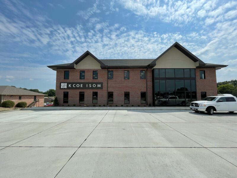 A brick office building with a sign saying "KCoe Isom" on the facade. A white pickup truck is parked in front, under a partly cloudy sky.