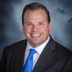 A smiling James Jirak, dressed in a suit with a blue tie, stands confidently against a cloudy backdrop.