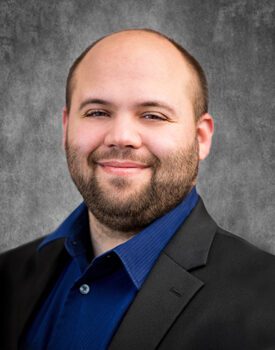 Joseph Brunner, sporting a beard and shaved head, is captured in a dark suit and blue shirt. He offers a slight smile against a gray textured background, exuding confidence and style.