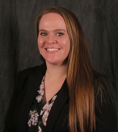 A person with long hair smiles, wearing a black blazer over a floral blouse, against a dark gray background.