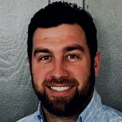 Ryan Green, a man with short dark hair and a beard, smiles at the camera in a light-colored collared shirt against a gray background.