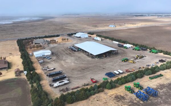 Aerial view of a large farmstead showcasing Food and Ag Production, with a metal barn, equipment, vehicles, trees lining the property, and wide fields surrounding the area under a partly cloudy sky.
