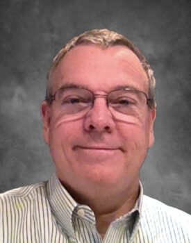A man in glasses and a striped shirt, who resembles Bill Penn, smiles warmly against a gray background.