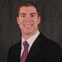 Trent Cooper is smiling in a suit and tie against a plain dark background.