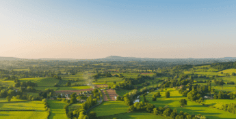 Aerial view of a lush green landscape with fields, trees, and a few houses under a clear blue sky, adhering to the GHG Protocol. Hills roll gently in the distance.