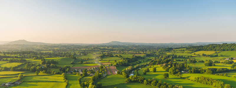 Aerial view of a lush green landscape with fields, trees, and a few houses under a clear blue sky, adhering to the GHG Protocol. Hills roll gently in the distance.