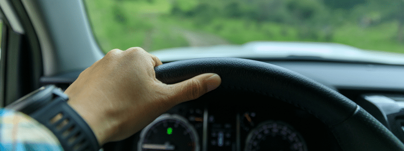 A hand grips the steering wheel of a car navigating a dirt road, green fields stretching wide in the background—a perfect escape from worries about auto depreciation limits.