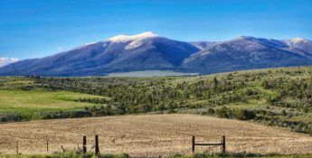 Experience the scenic view of a vast landscape featuring grassy fields, a conservation easement with fenced areas, and snow-peaked mountains under a clear blue sky.